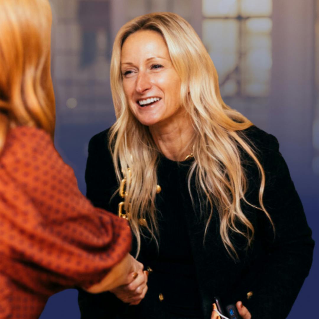 A blonde, smiling woman in a black jacket shaking hands with a woman in an orange blouse who is facing away from the camera