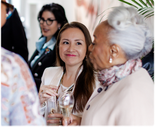 A group of women holding drinks and engaging in conversation with the camera focused on one woman listening and smiling